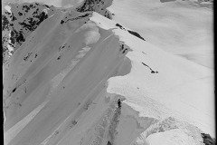 Blick auf verschneite Berggipfel, inmitten zwei Skifahrer, die ihre Skier tragen und den Berg herablaufen, Feld mit Skispuren durchzogen, dahinter der bewölkte Himmel